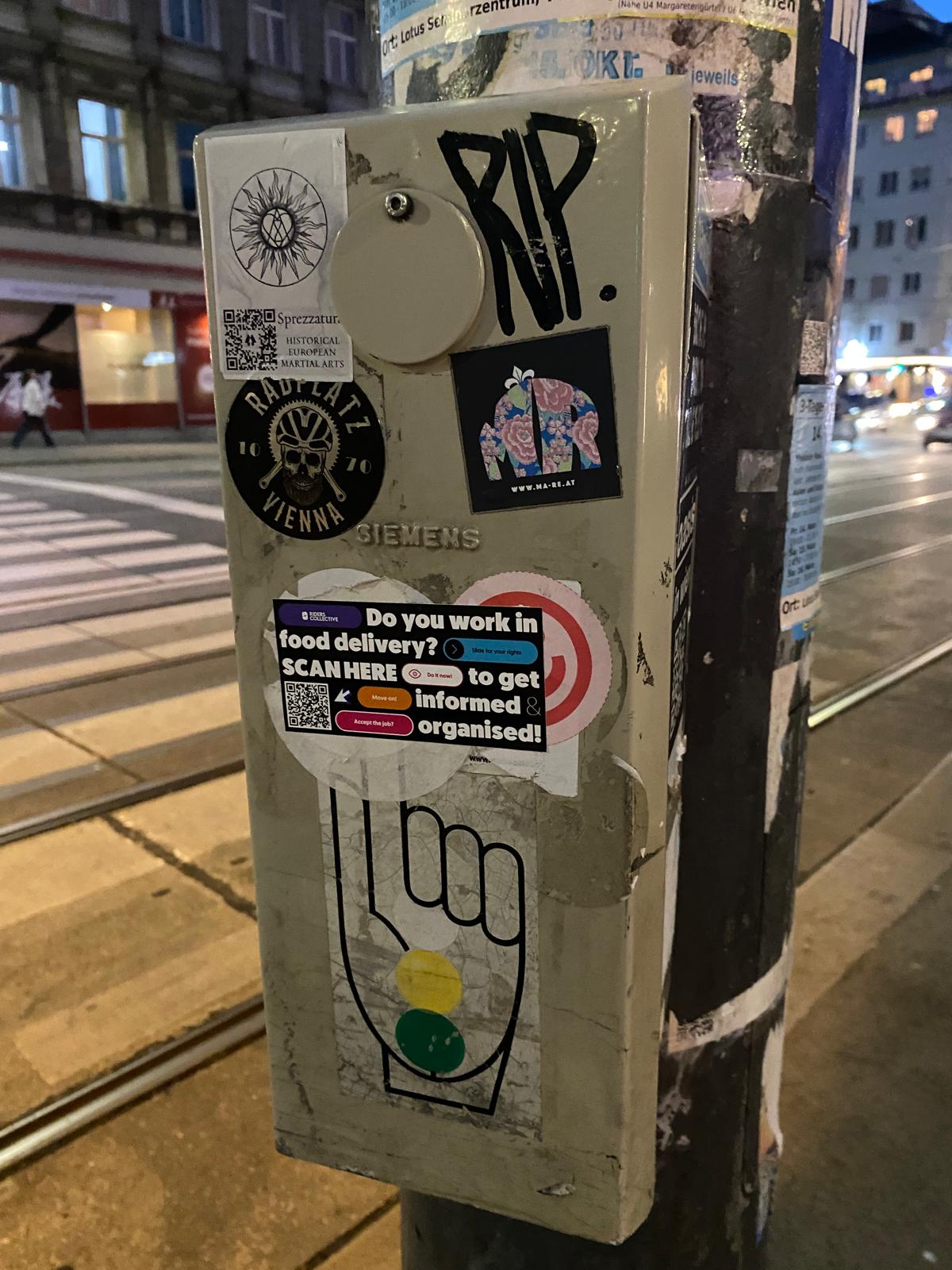 Mehrere Aufkleber auf einem Elektrik-Kasten an einer Ampelanlage. Auf einem Sticker steht: Do you work in food delivery? Scan here to get informed & orgnaised!